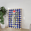 Decorative wall art with a blue and white checkered border and floral design in a room with a plant and vase.