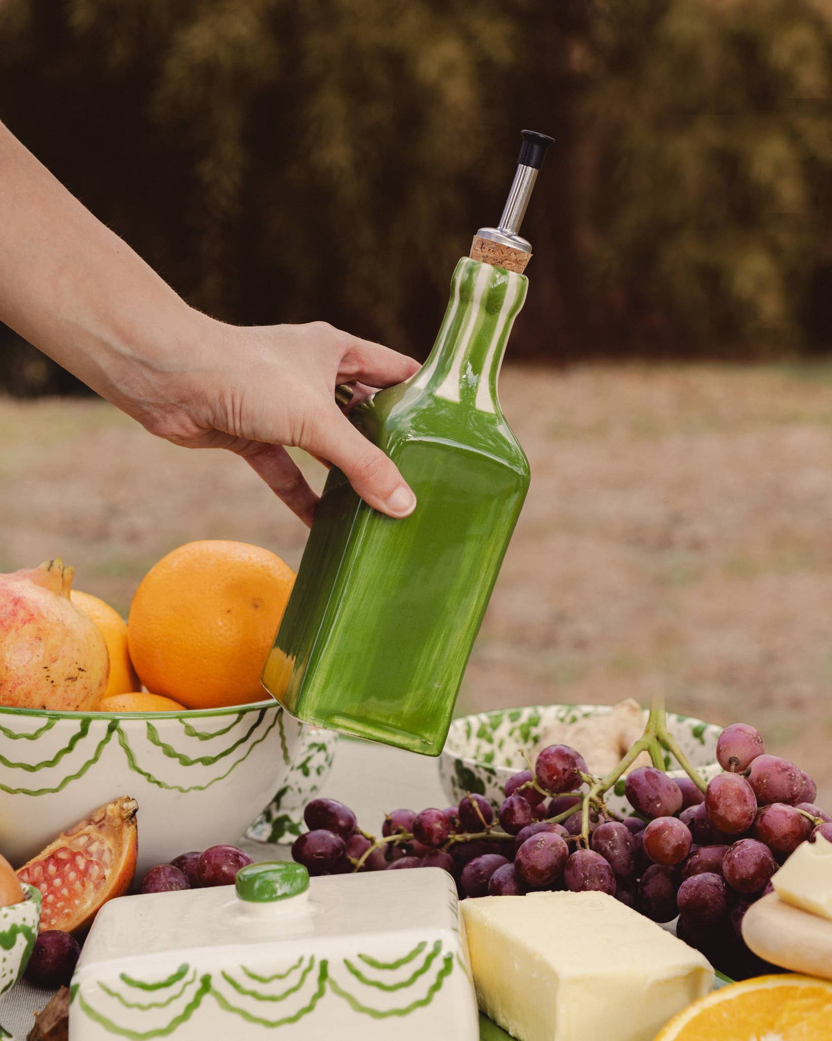 Person holding a ceramic green oil pourer with fruits and cheese on a table outdoors.