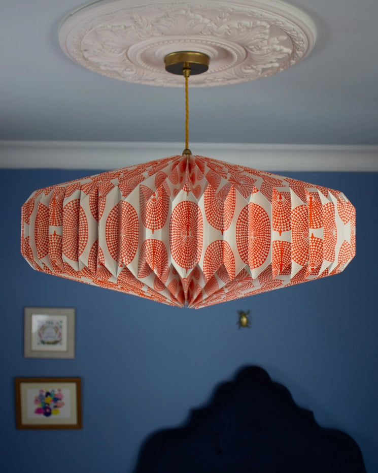 A modern octagonal pendant light with a white and coral-orange geometric pattern, hanging from a brass fixture in a white decorative ceiling medallion against a blue wall.