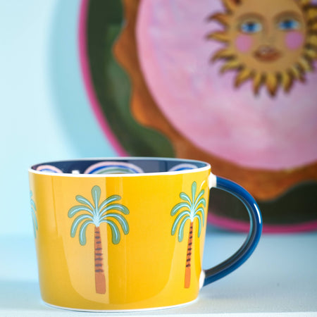Yellow mug with palm tree design in front of a decorative plate with a sun face.