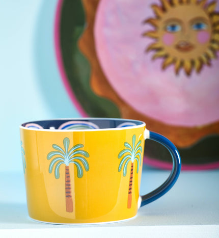 Yellow mug with palm tree design in front of a decorative plate with a sun face.