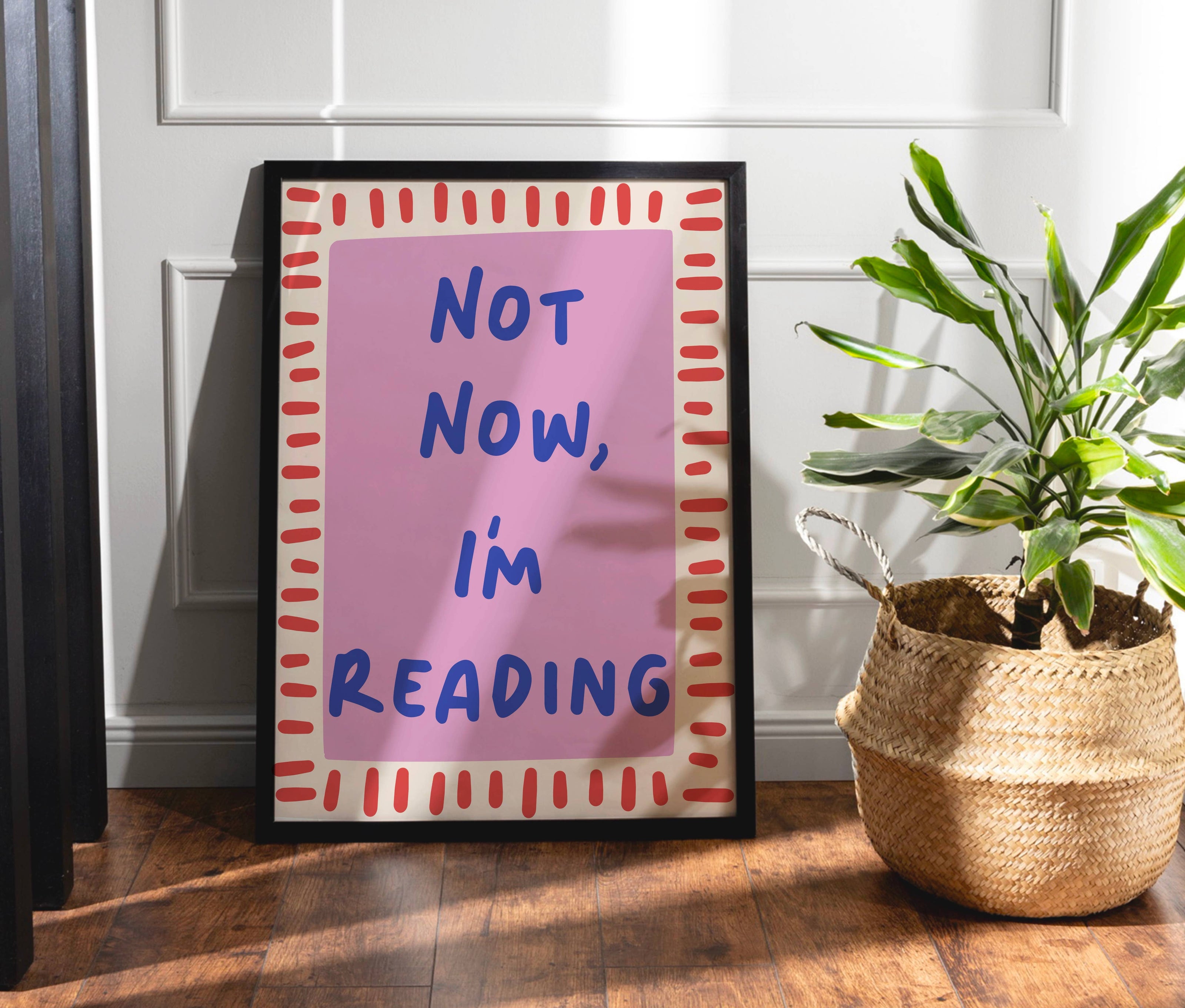 Framed poster with 'Not Now, I'm Reading' text next to a plant on a wooden floor.