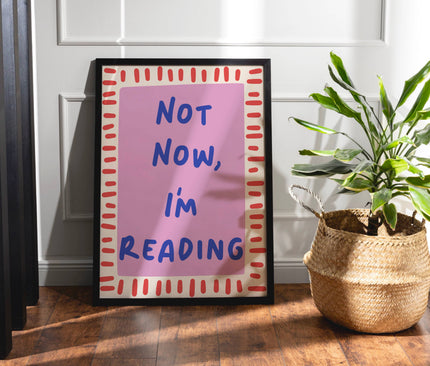 Framed poster with 'Not Now, I'm Reading' text next to a plant on a wooden floor.