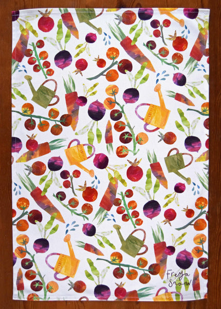 Colorful garden-themed tea towel with vegetables and watering cans on a white background.