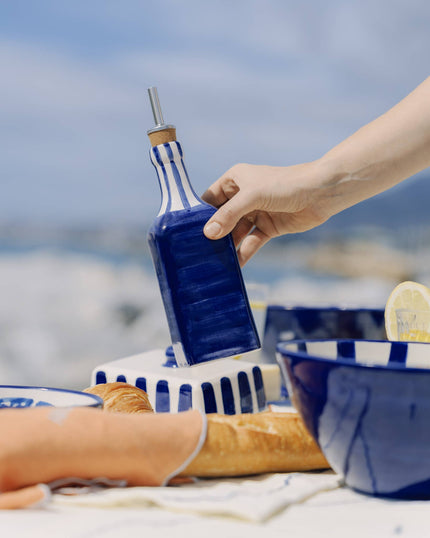 Hand holding a blue ceramic oil pourer set against a blurred outdoor background.