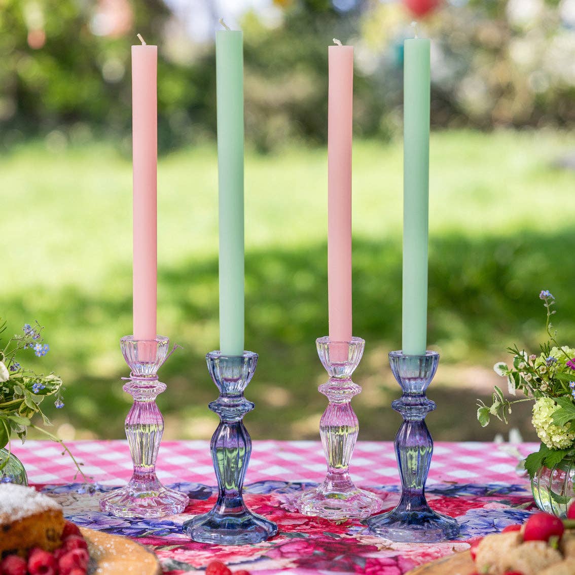 Four pastel-colored candles in decorative candlesticks on a table outdoors.
