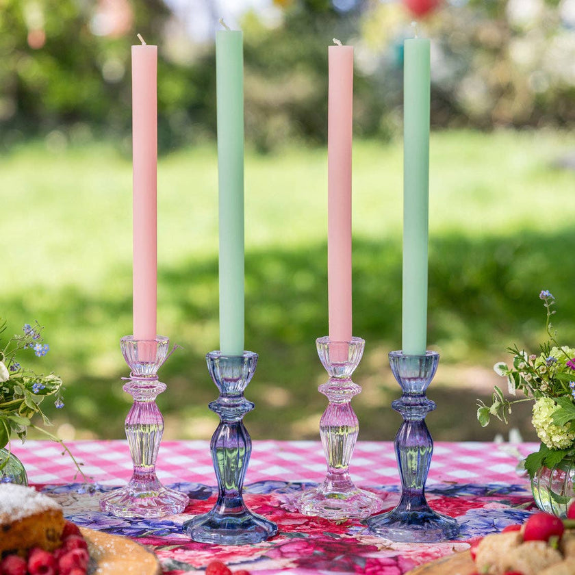 Four pastel-colored candles in decorative candlesticks on a table outdoors.