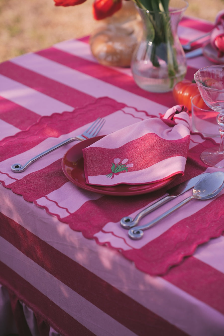 Table setting with red and white striped tablecloth, pink napkin, and silverware on a blurred outdoor background.