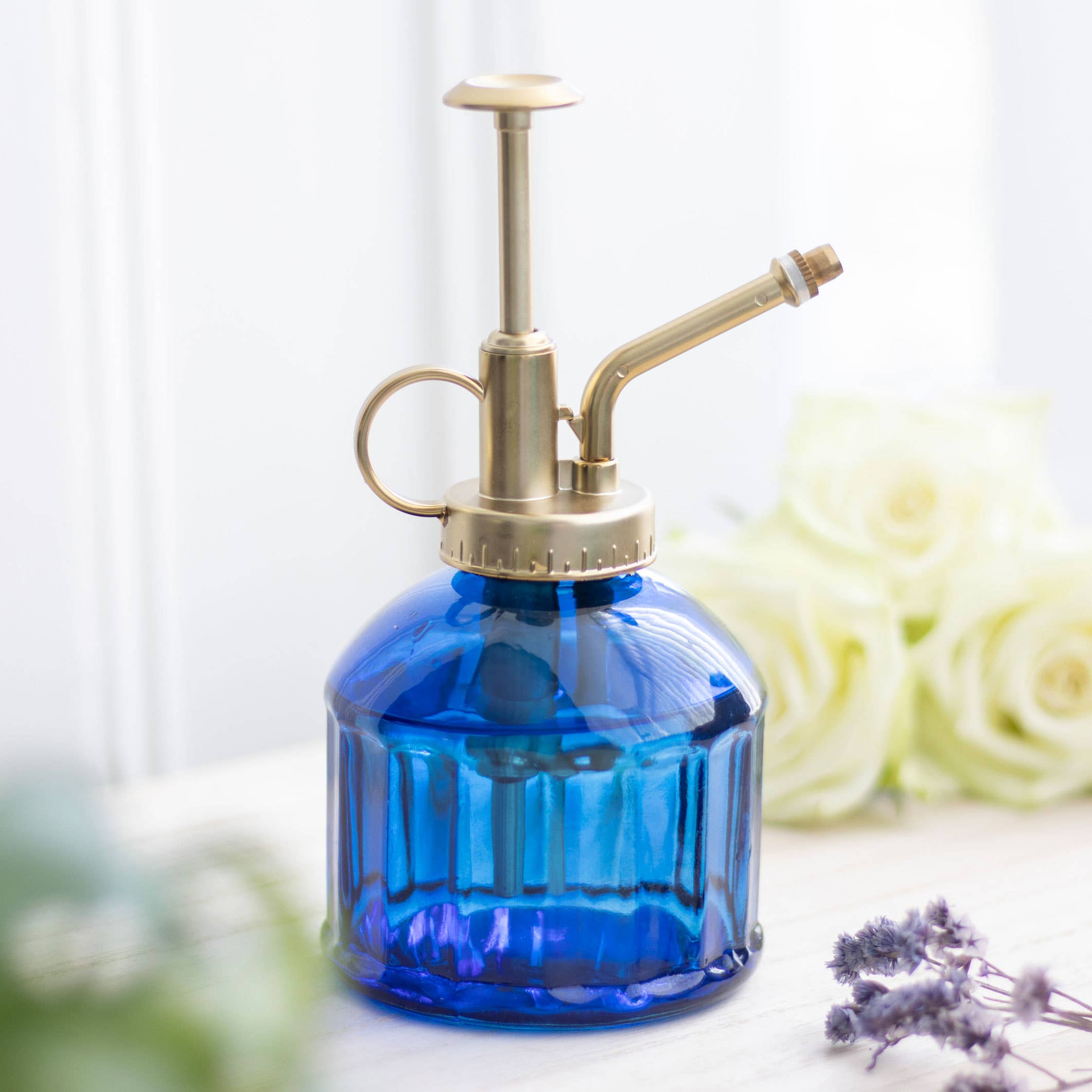 Blue glass spray bottle with brass nozzle on a white surface with flowers in the background
