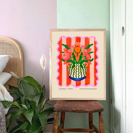 Framed artwork of a floral vase on a wooden stool with plants and a chair in the background.