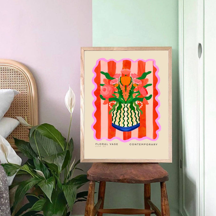 Framed artwork of a floral vase on a wooden stool with plants and a chair in the background.