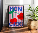 Framed artwork with 'MON çHERI' text and cherries on a striped background, placed on a wooden floor.