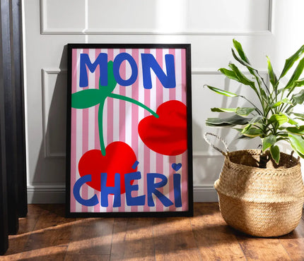 Framed artwork with 'MON çHERI' text and cherries on a striped background, placed on a wooden floor.