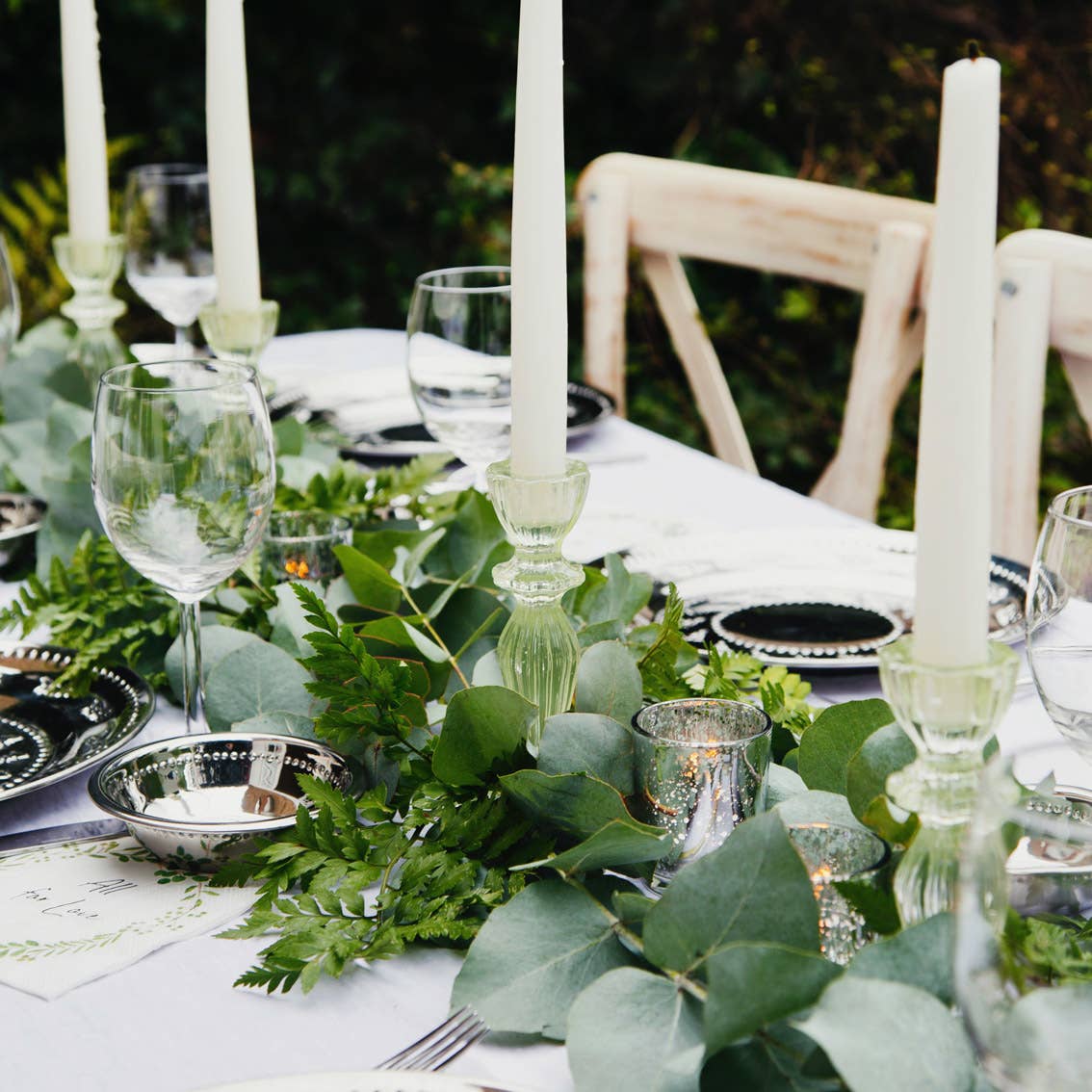Elegant table setting with greenery, candles, and glassware on a white tablecloth.